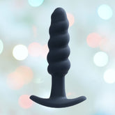 The VeDO Twist Plug Vibrating Butt Plug in black, featuring a beaded design and 10 vibration modes, is displayed against a blurred background with soft, pastel-colored lights.